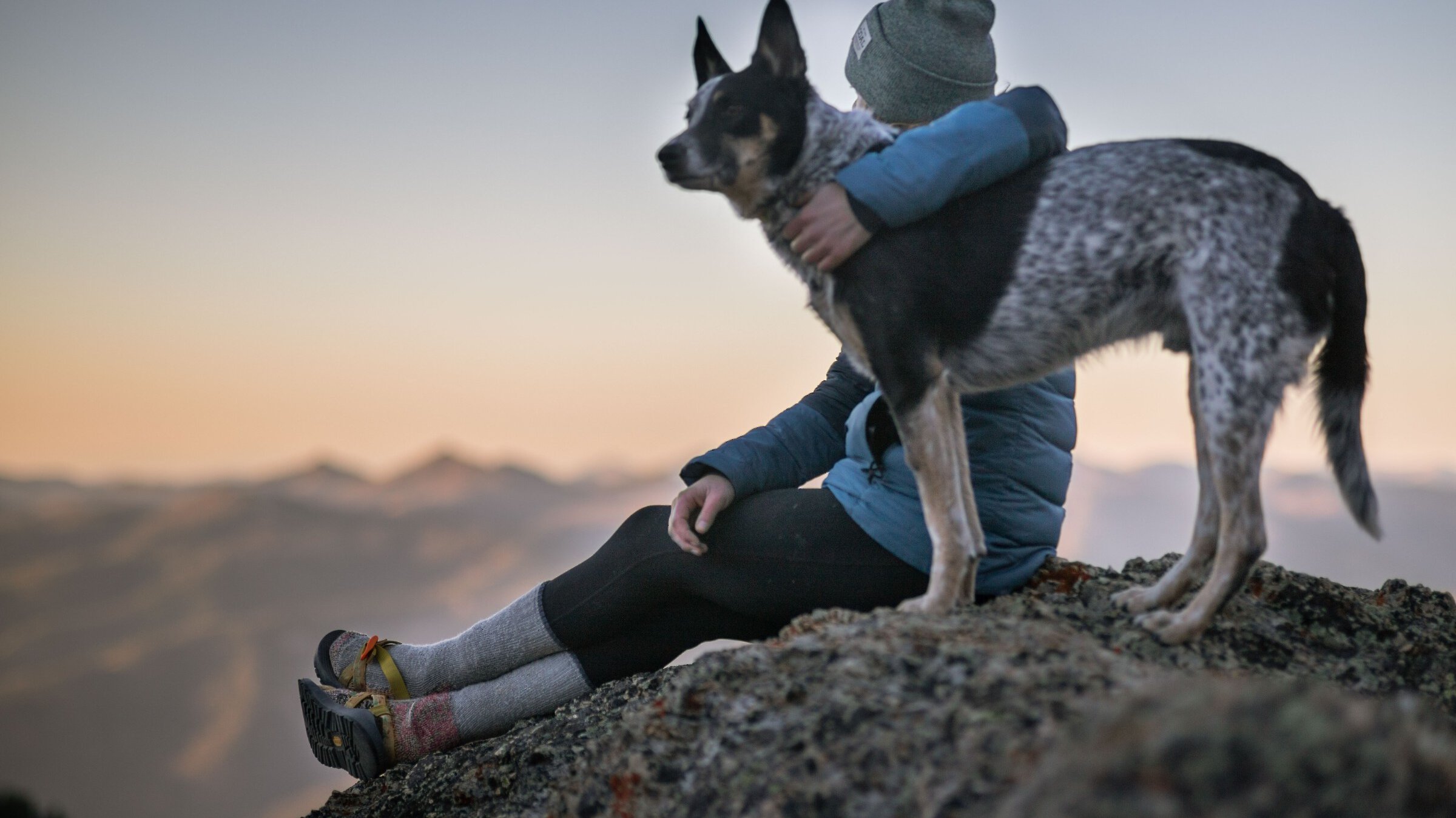 Woman with arm around dog while sitting on a mountain top at sunset